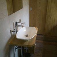 Nowoczesna, owalna umywalka nablatowa z chromowaną baterią zamontowana na drewnianym blacie w łazience wykończonej płytkami w odcieniach beżu i brązu. Widoczne odbicie w lustrze i fragment drzwi.