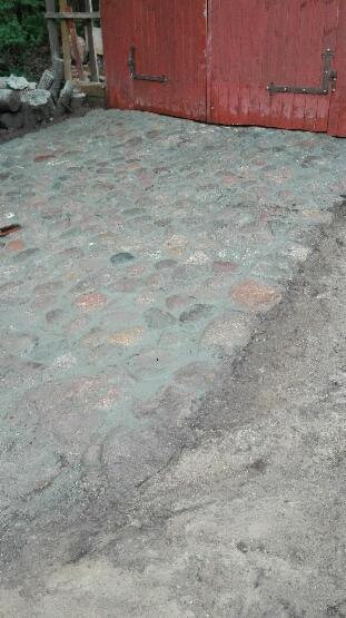 Ułożona z kamieni ścieżka lub podjazd z widocznymi spoinami, przylegająca do czerwonych drewnianych drzwi i fragmentu kamiennego murku.