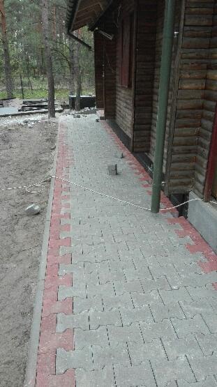 Ułożona szara kostka brukowa z czerwonym obramowaniem przy drewnianym domu, widoczne niedokończone prace i piasek.