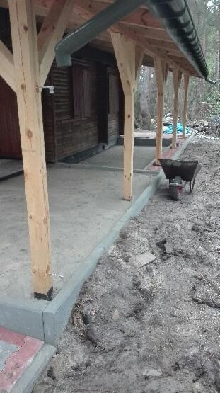 Zadaszony taras z surowego drewna przy domku letniskowym, świeżo wylana betonowa wylewka, widoczna taczka z czerwoną ramą i czarną skrzynią.