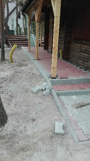 Układanie chodnika z kostki brukowej przy drewnianym budynku z kolumnami, widoczne niedokończone fragmenty i piasek.