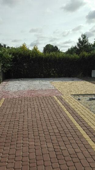 Ułożona kostka brukowa w odcieniach brązu i żółci, z fragmentami w kolorze białym i czerwonym, otoczona zielenią, w tle pochmurne niebo.