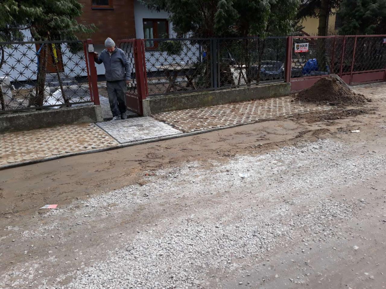 Układanie ażurowych płyt chodnikowych przed posesją, widoczny mężczyzna wchodzący przez furtkę, obok sterta ziemi.