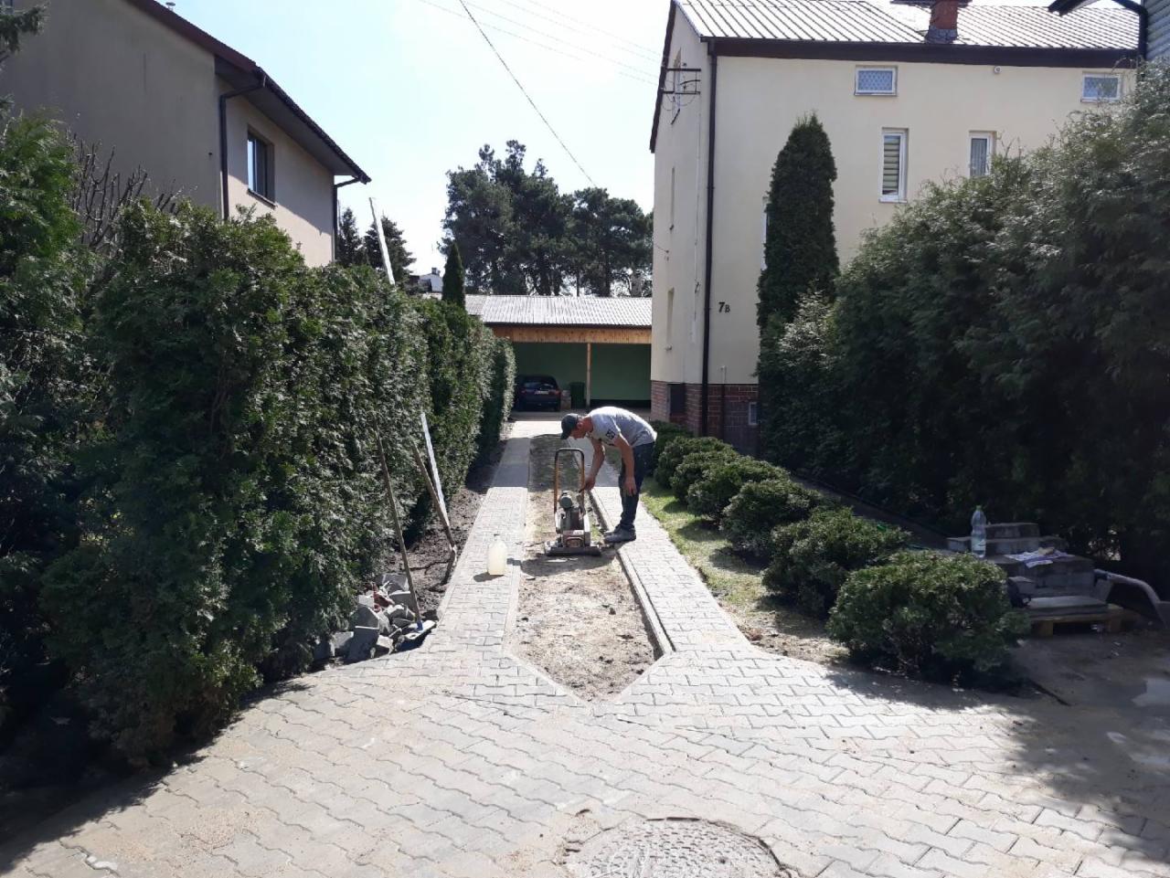 Pracownik zagęszcza podłoże pod kostkę brukową na podjeździe do domu przy użyciu zagęszczarki spalinowej, widoczne przygotowane kostki i obrzeża.
