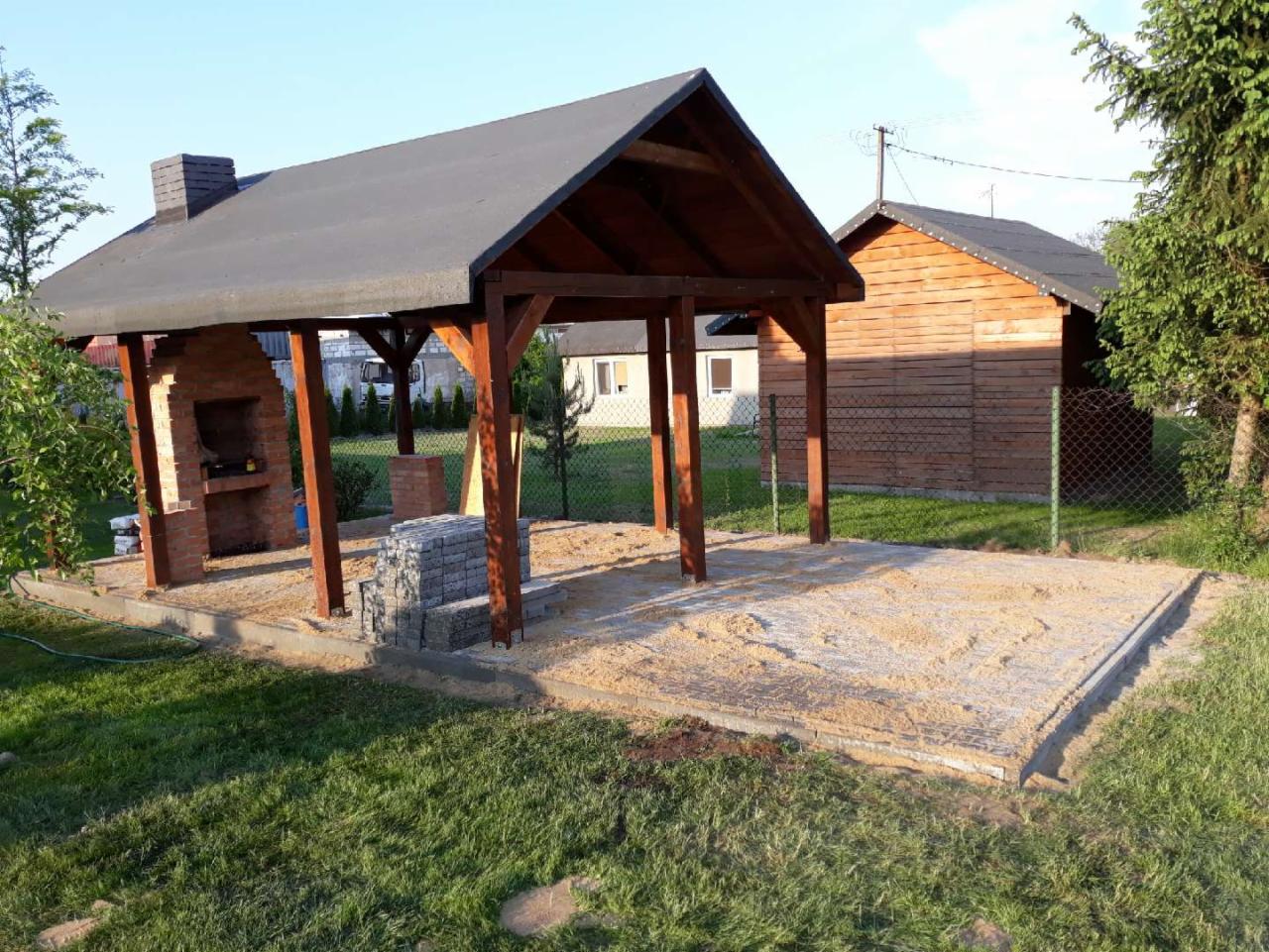 Budowa zadaszonej przestrzeni grillowej z ceglanym paleniskiem i drewnianą konstrukcją dachu, widoczne przygotowane podłoże z piasku i kostki do wykończenia.