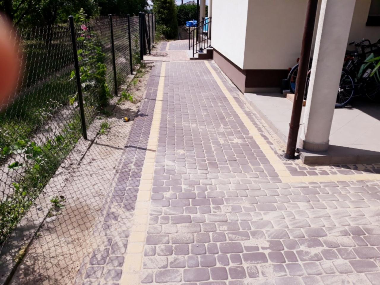 Ułożona kostka brukowa w odcieniach szarości i beżu tworząca ścieżkę obok domu, widoczny fragment ogrodzenia z siatki i roślinność.