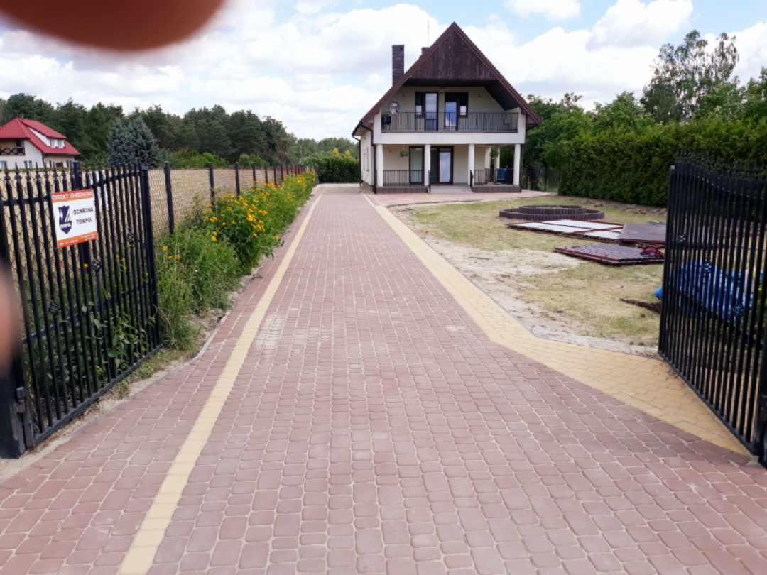 Ułożona z kostki brukowej, ceglastoczerwona ścieżka prowadząca do dwukondygnacyjnego domu z balkonem, widok zza otwartej, czarnej, metalowej bramy.