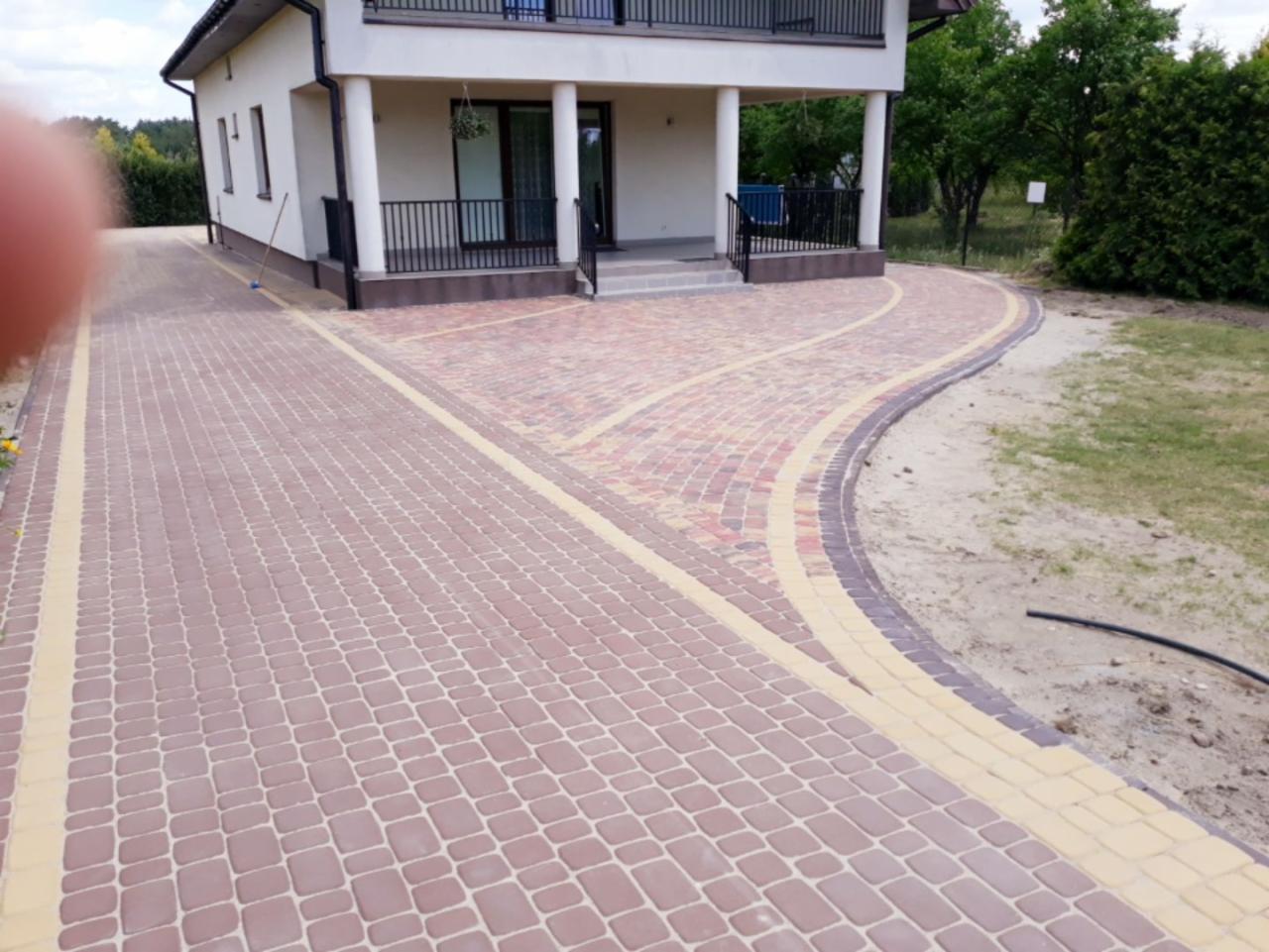 Ułożona kostka brukowa w odcieniach brązu i beżu tworząca podjazd do domu z białymi kolumnami i czarną balustradą.