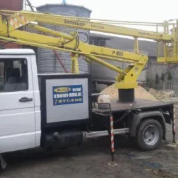 Żółty podnośnik koszowy zamontowany na białym samochodzie dostawczym z reklamą firmy, na ziemi leżą betonowe bloczki, w tle silosy i budynki.