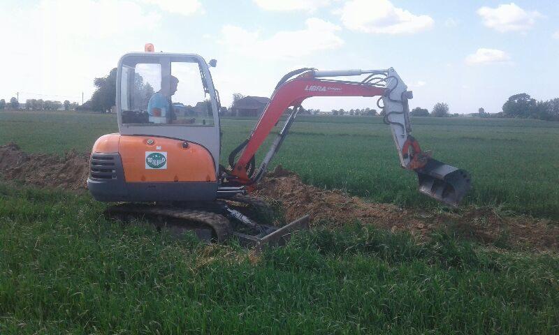 Pomarańczowa minikoparka Libra z operatorem w kabinie, kopiąca rów na polu z zieloną trawą, widoczne świeżo wykopane grudki ziemi.