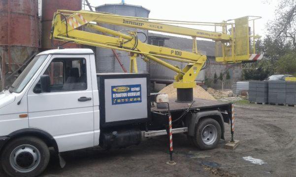 Żółty podnośnik koszowy zamontowany na białym samochodzie dostawczym z reklamą firmy, na ziemi leżą betonowe bloczki, w tle silosy i budynki.