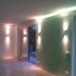 Salon z zieloną ścianą akcentową oświetloną przez cztery nowoczesne kinkiety ścienne, dodatkowo pod sufitem zainstalowano listwę LED z zielonym światłem, widoczne duże okna balkonowe.