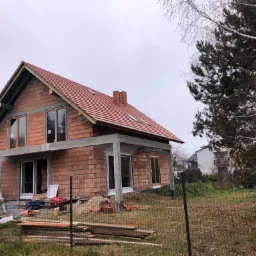 Niedokończony dom murowany z czerwoną dach&oacute;wką, widoczne okna bez wykończenia, betonowe nadproża, na placu budowy leżą deski i stoi agregat prądotw&oacute;rczy.