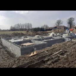 Wylane fundamenty z bloczk&oacute;w betonowych na błotnistym terenie budowy, widoczne drewniane elementy szalunku i stosy bloczk&oacute;w w tle, pochmurne niebo.