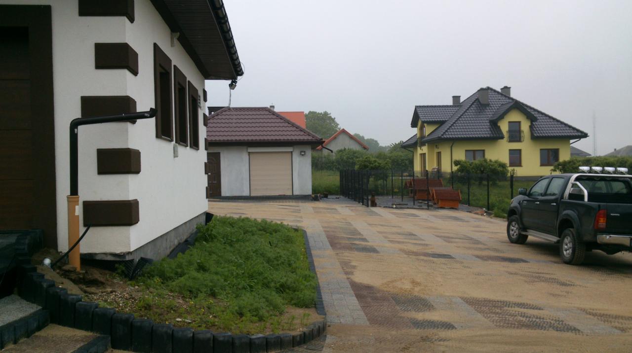 Ułożona kostka brukowa na podjeździe z widocznymi śladami opon samochodu terenowego, w tle budynki mieszkalne w pochmurny dzień.