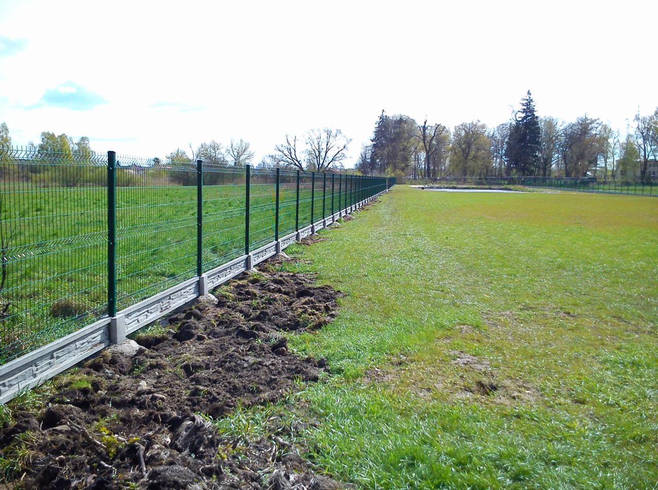 Długi, zielony płot z siatki panelowej na betonowej podmurówce z dekoracyjnym wzorem, oddzielający trawnik od pola, widoczne świeżo przekopane fragmenty ziemi przy fundamencie.
