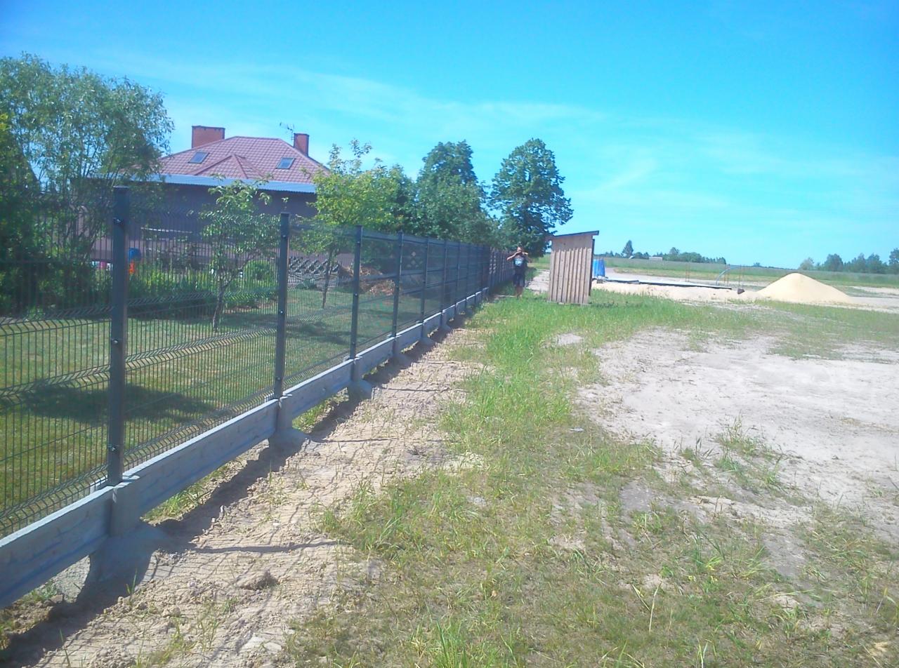 Nowo postawione ogrodzenie panelowe z podmurówką betonową na działce z widocznym domem i stertą piasku w tle.