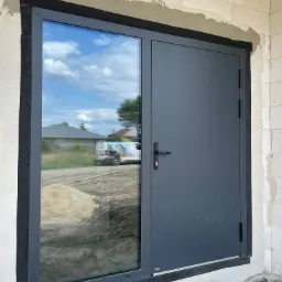 Montaż okna aluminiowego dwuskrzydłowego w surowym stanie budynku, widoczne uszczelnienie i odbicie krajobrazu w szybie.