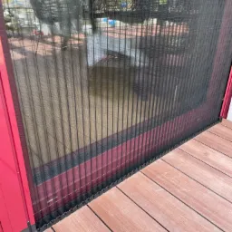 Zewnętrzna moskitiera plisowana zamontowana na czerwonej ramie drzwiowej, widok z bliska na taras z desek kompozytowych.