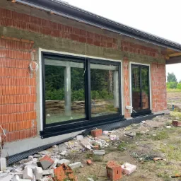 Surowy stan budynku w trakcie montażu okien z czarnymi ramami; widoczne niedokończone ściany z cegły, wystające kable i gruz budowlany na zewnątrz.