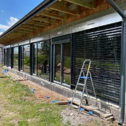 Montaż żaluzji fasadowych zewnętrznych na budynku w stanie surowym, widoczna drabina, narzędzia i pracownik przy oknie, ściany z pustaków ceramicznych.