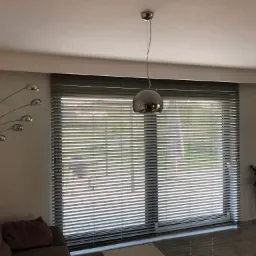 Szare żaluzje poziome zamontowane w oknie w Tomaszowie Mazowieckim, widoczne z wnętrza pokoju, z lampą wiszącą i dekoracyjną lampą podłogową.