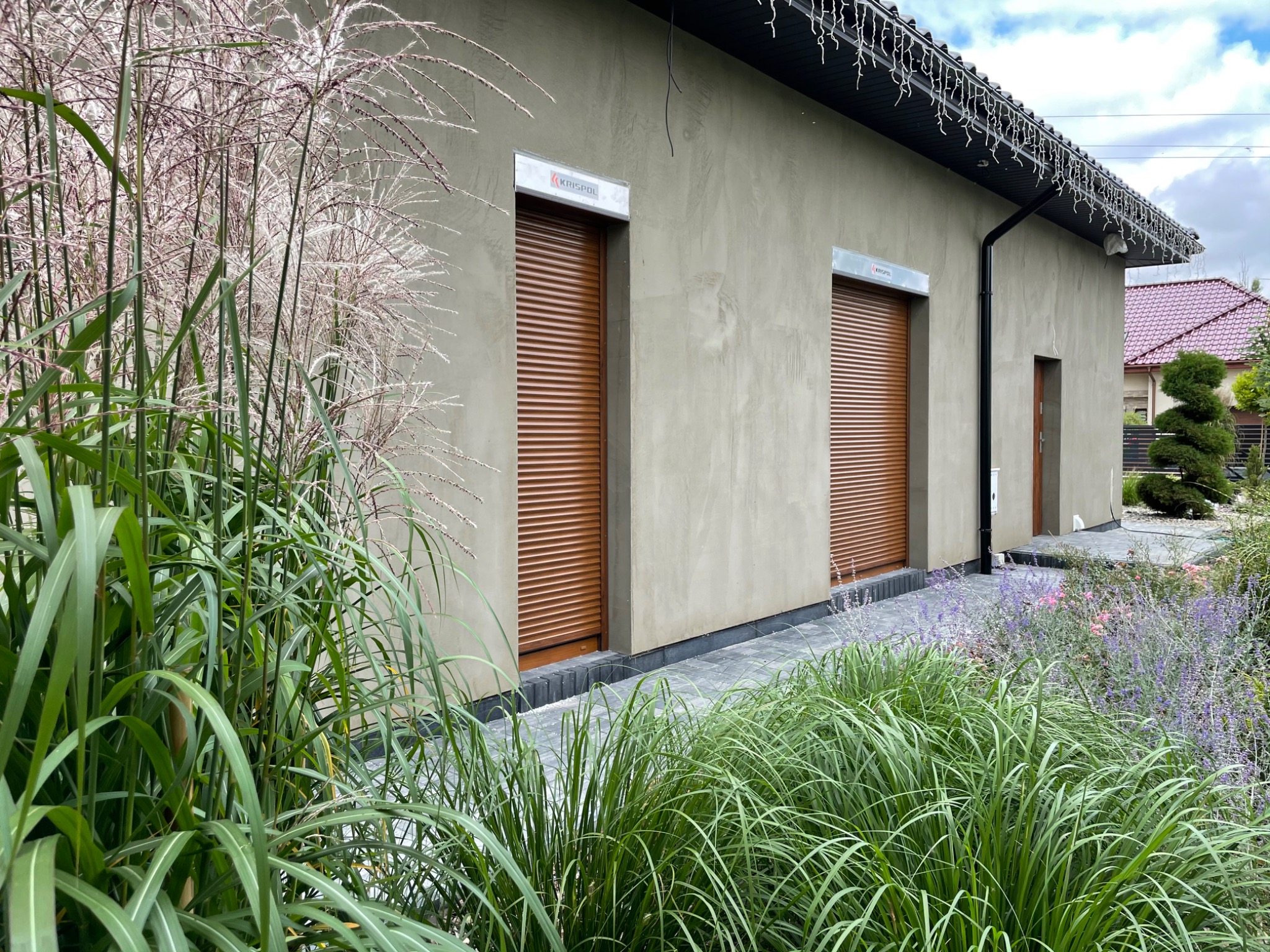 Budynek gospodarczy z dwoma brązowymi roletami zewnętrznymi, widoczne logo Krispol nad jedną z nich, otoczony dekoracyjną roślinnością.