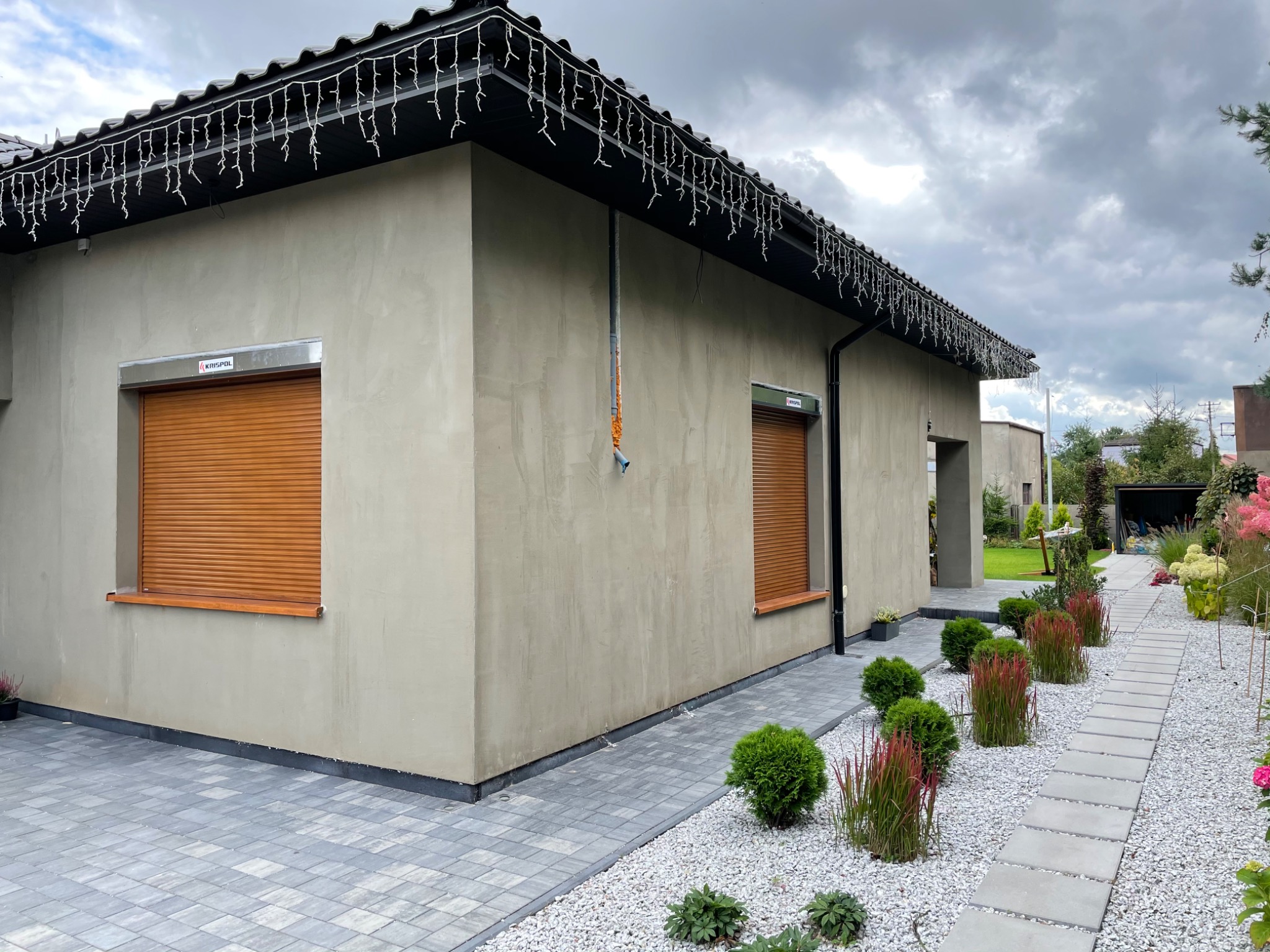 Dom jednorodzinny z beżową elewacją i brązowymi roletami zwiniętymi na oknach, oświetlenie świąteczne pod dachem, ścieżka z płyt betonowych w białym żwirze.