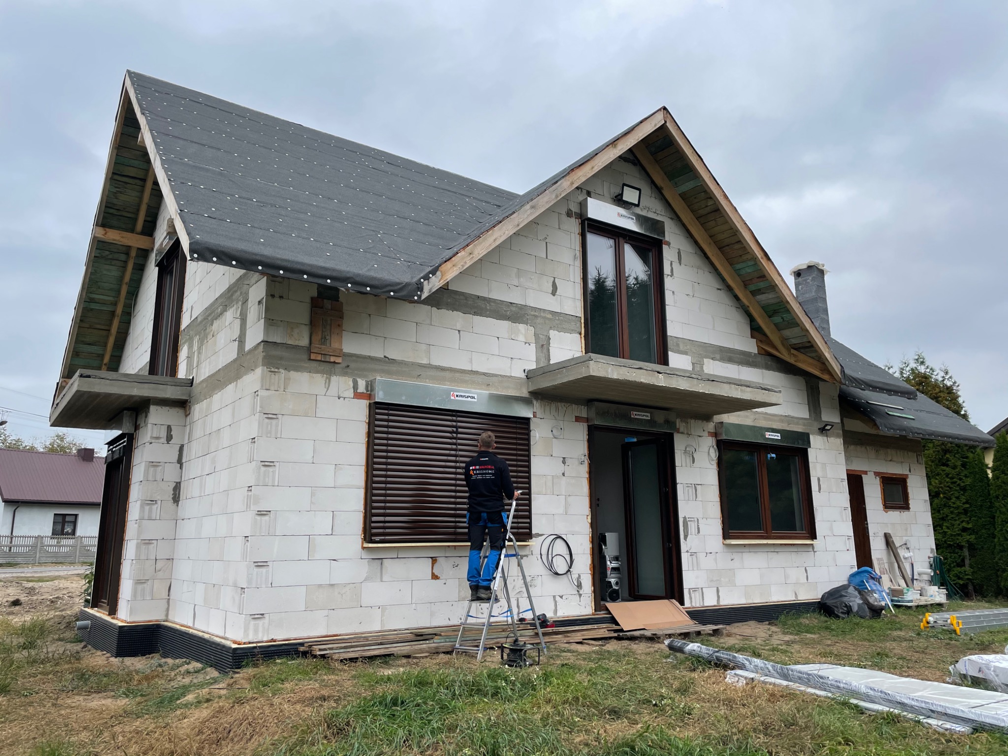 Montaż brązowych rolet zewnętrznych na surowym budynku z białego bloczka, pracownik na drabinie, widoczne materiały budowlane i narzędzia.