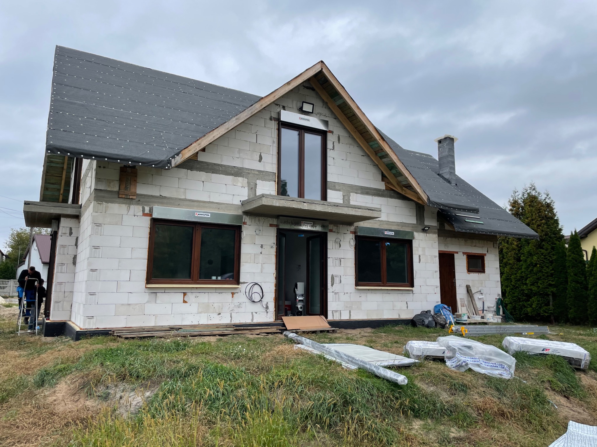 Surowy stan nowo budowanego domu z zamontowanymi oknami z brązowymi ramami i częściowo zamontowanymi roletami zewnętrznymi, widoczne materiały budowlane i drabina.