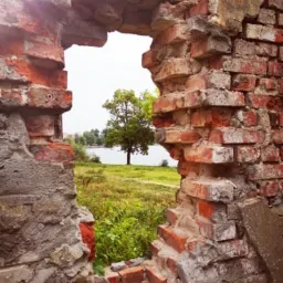 Zrujnowana ceglana ściana z widokiem na jezioro i drzewo przez otwór. Widoczne ślady erozji i brakujące fragmenty cegieł.