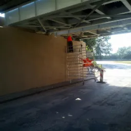 Pracownik w pomarańczowej kamizelce na rusztowaniu przy ścianie wiaduktu, na której widać świeżo nałożoną warstwę izolacyjną, z widocznymi śladami po narzędziach.