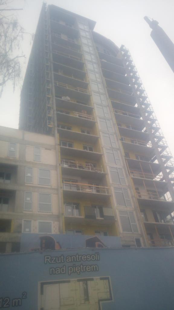 Wysoki budynek w trakcie budowy, widoczne rusztowania, niezabezpieczone balkony i baner reklamowy z rzutem antresoli.