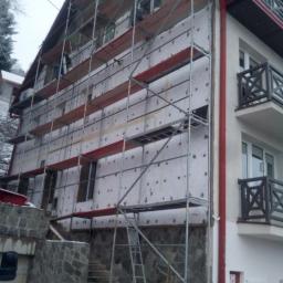 drew-nex - Ocieplanie elewacji budynku wielorodzinnego z użyciem rusztowania, widoczne płyty izolacyjne z szarymi kołkami mocującymi, częściowo wykończona elewacja, kamienne schody wejściowe.