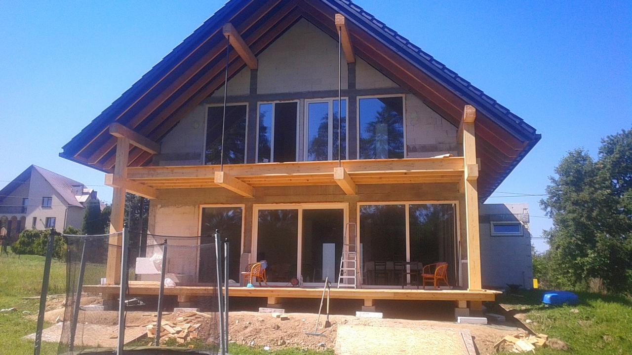 Surowy stan dwukondygnacyjnego domu z drewnianą konstrukcją szkieletową i dużymi oknami, widoczne drewniane elementy konstrukcyjne dachu i tarasu, ziemia wokół domu częściowo uprzątnięta, w tle...