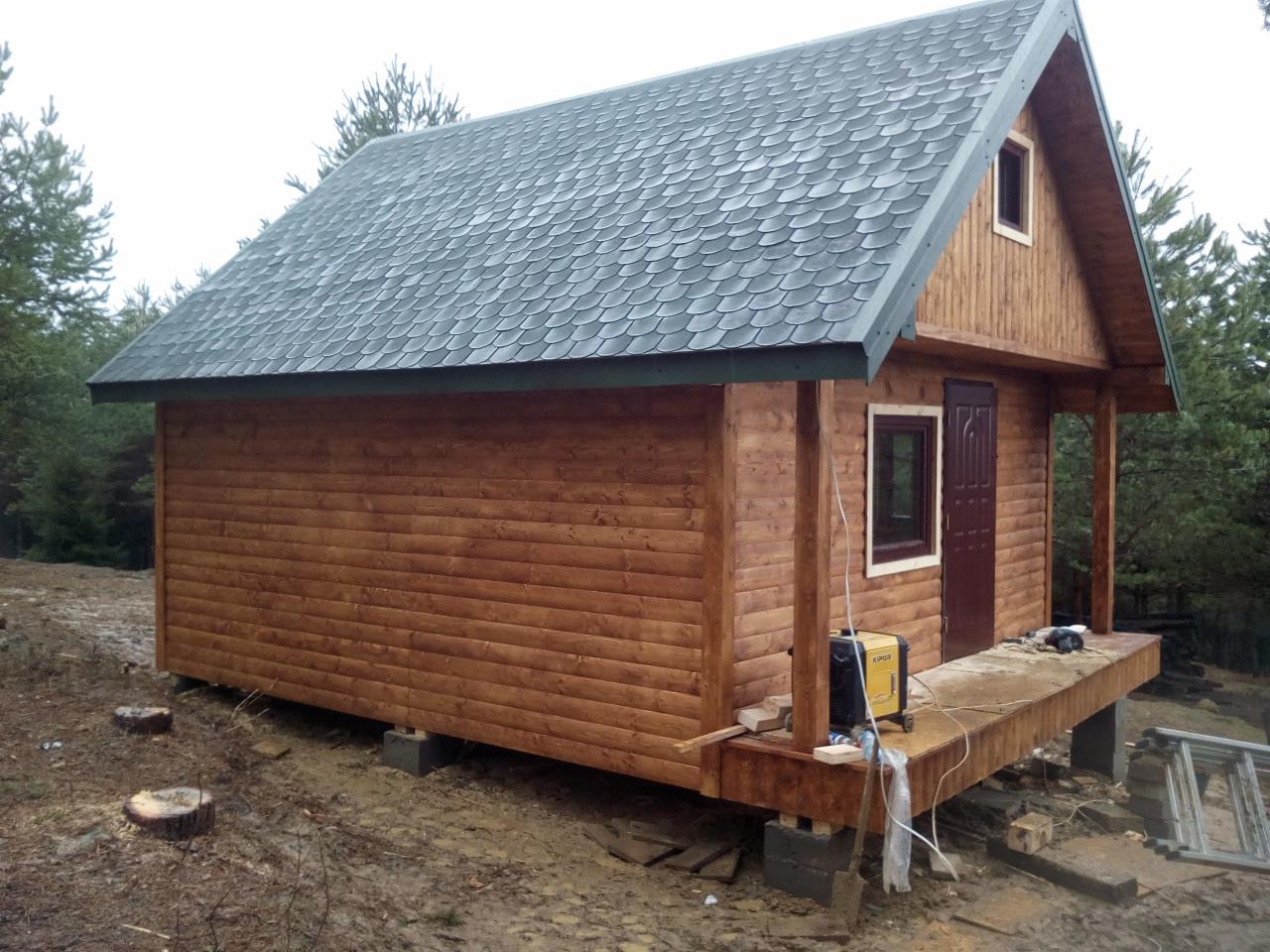 Niedokończony drewniany domek z ciemnym dachem z gontu bitumicznego, widoczny agregat prądotwórczy i drabina, teren budowy.