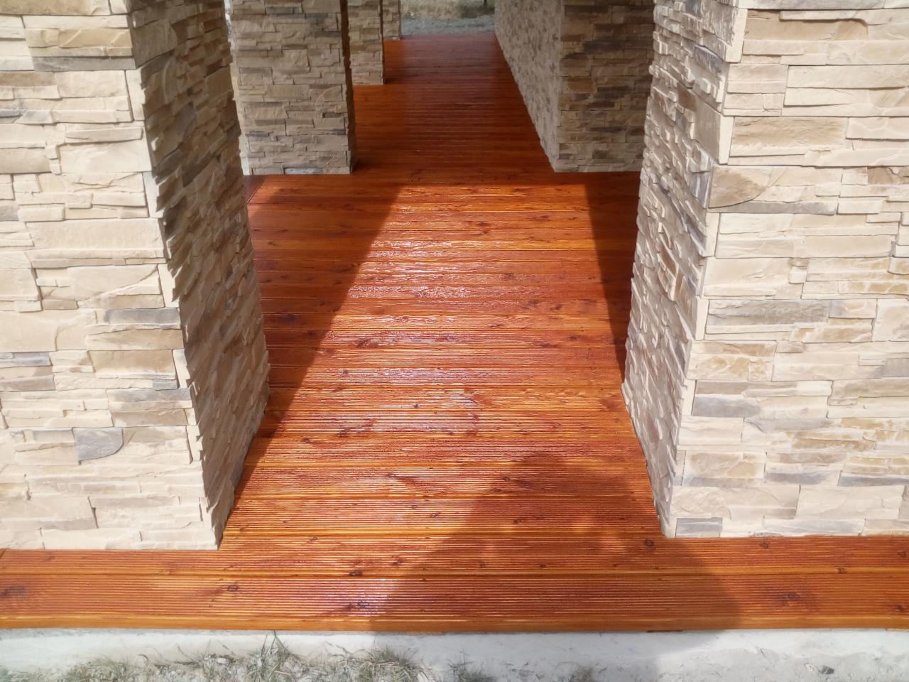 Zadaszone przejście z drewnianą podłogą z desek i kamiennymi filarami w odcieniach beżu, widoczne refleksy światła na lakierowanej powierzchni podłogi.