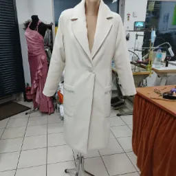 Elegancki, kremowy płaszcz damski z ozdobnymi paskami cyrkonii przy kieszeniach, prezentowany na manekinie w pracowni krawieckiej, z widoczną suknią wieczorową na drugim manekinie w tle.