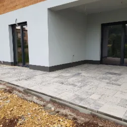 Jasnoszara kostka brukowa ułożona na tarasie przed nowoczesnym budynkiem z dużymi przesuwnymi drzwiami, widoczne przygotowane podłoże z kamieniami.