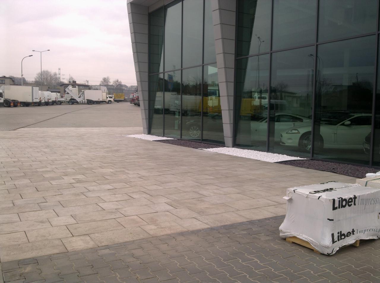 Nowoczesna kostka brukowa przed biurowcem z paletą kostki Libet Impression w rogu, białe kamienie ozdobne przy elewacji, parking z ciężarówkami w tle.