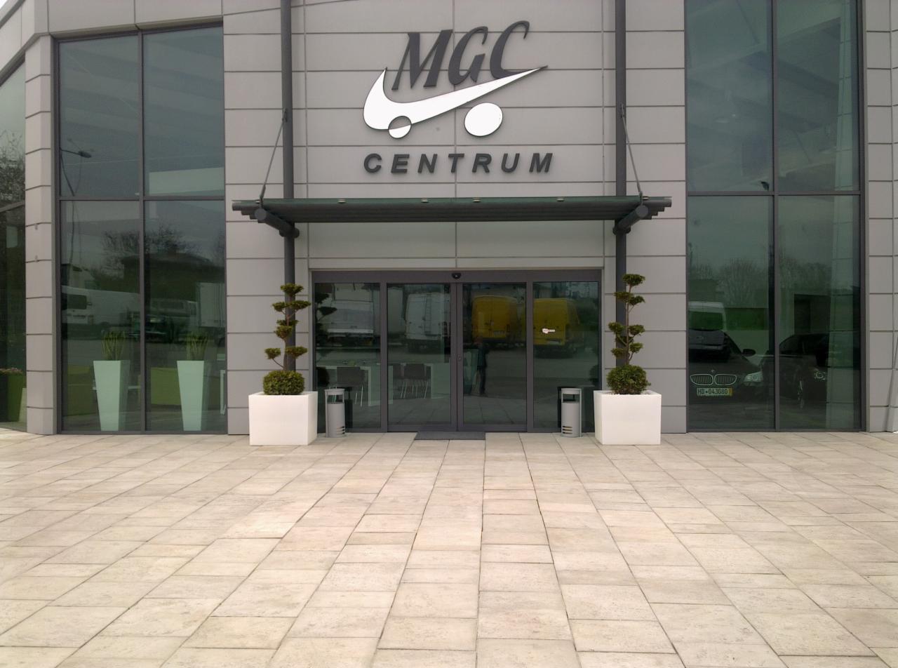 Jasna kostka brukowa przed nowoczesnym budynkiem biurowym z dużymi oknami i logo MGC Centrum, ozdobne drzewka w białych donicach po bokach wejścia.