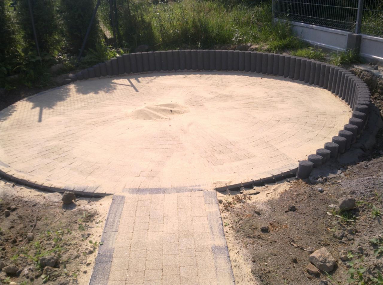 Przygotowany teren pod okrągły taras z kostki brukowej, otoczony palisadą, z widoczną ścieżką wejściową, etap prac ziemnych i podsypki piaskowej.