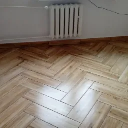 Ułożona w jodełkę podłoga z płytek imitujących drewno, z widocznym grzejnikiem i fragmentem białej ściany z listwą przypodłogową w kolorze płytek.