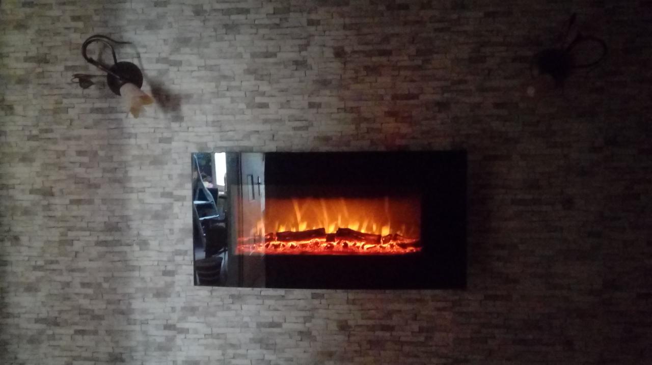 Ściana z dekoracyjnej cegły z elektrycznym kominkiem w czarnej obudowie i dwoma kinkietami po bokach.