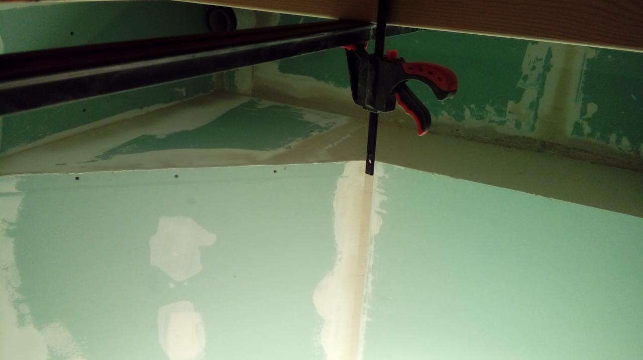 Mocowanie płyty karton-gips do sufitu za pomocą zacisku stolarskiego, widoczne szpachlowane łączenia płyt i fragment drewnianej konstrukcji stropu.