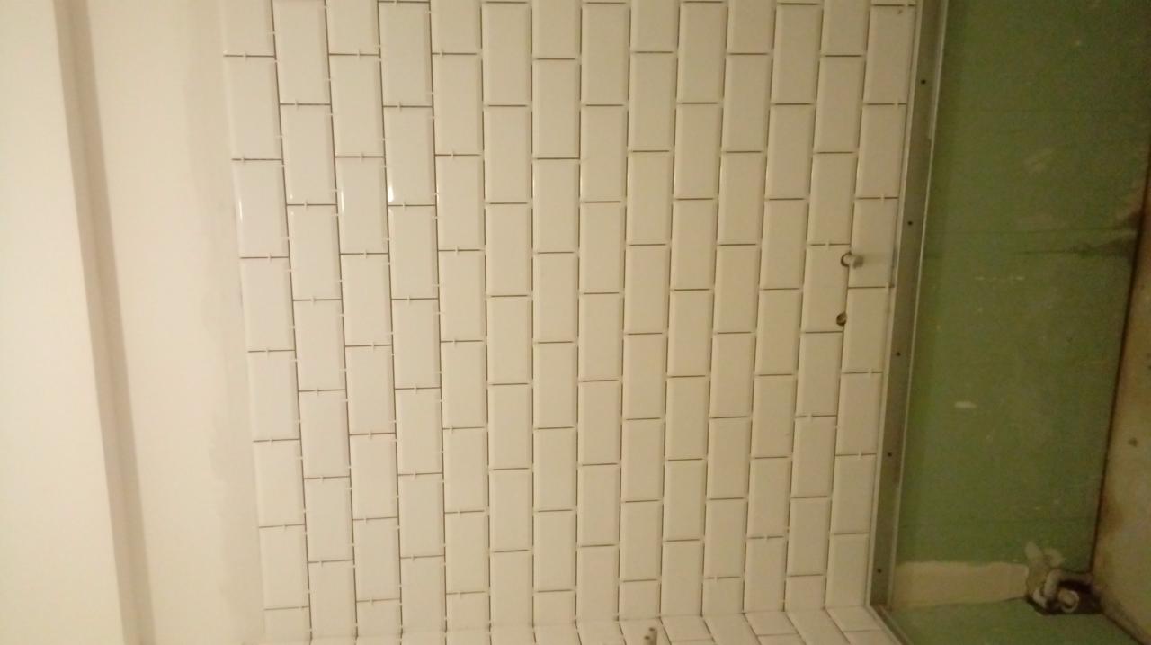 Ściana łazienki w trakcie układania białych, prostokątnych płytek, widoczne niedociągnięcia i surowy stan ściany obok.