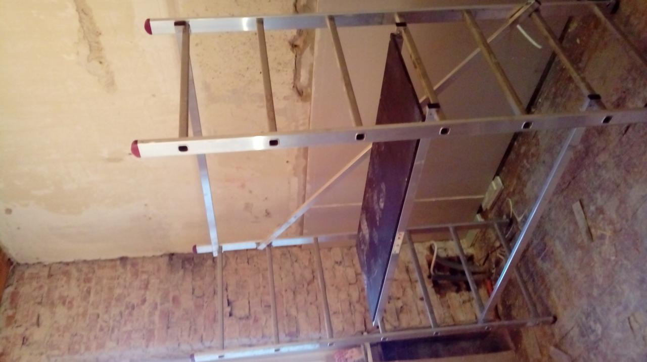 Metalowy ruszt ze stopniami na tle ściany z widocznym fragmentem cegieł i fragmentem sufitu w trakcie prac wykończeniowych. Widoczne ślady po usuniętych płytkach lub gładzi.