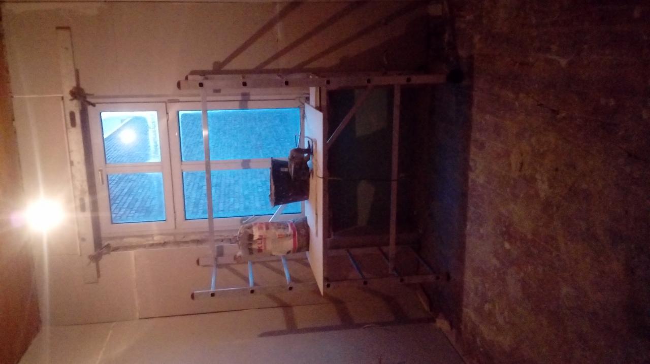 Wnętrze w trakcie prac wykończeniowych: ściany częściowo pokryte płytami, widoczny rusztowanie, okno z widokiem na zewnątrz, pojemnik z zaprawą. Oświetlenie punktowe.