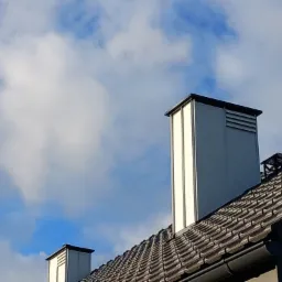 Nowoczesny dom z dachówką ceramiczną i metalowymi kominami wentylacyjnymi na tle błękitnego nieba z białymi chmurami.