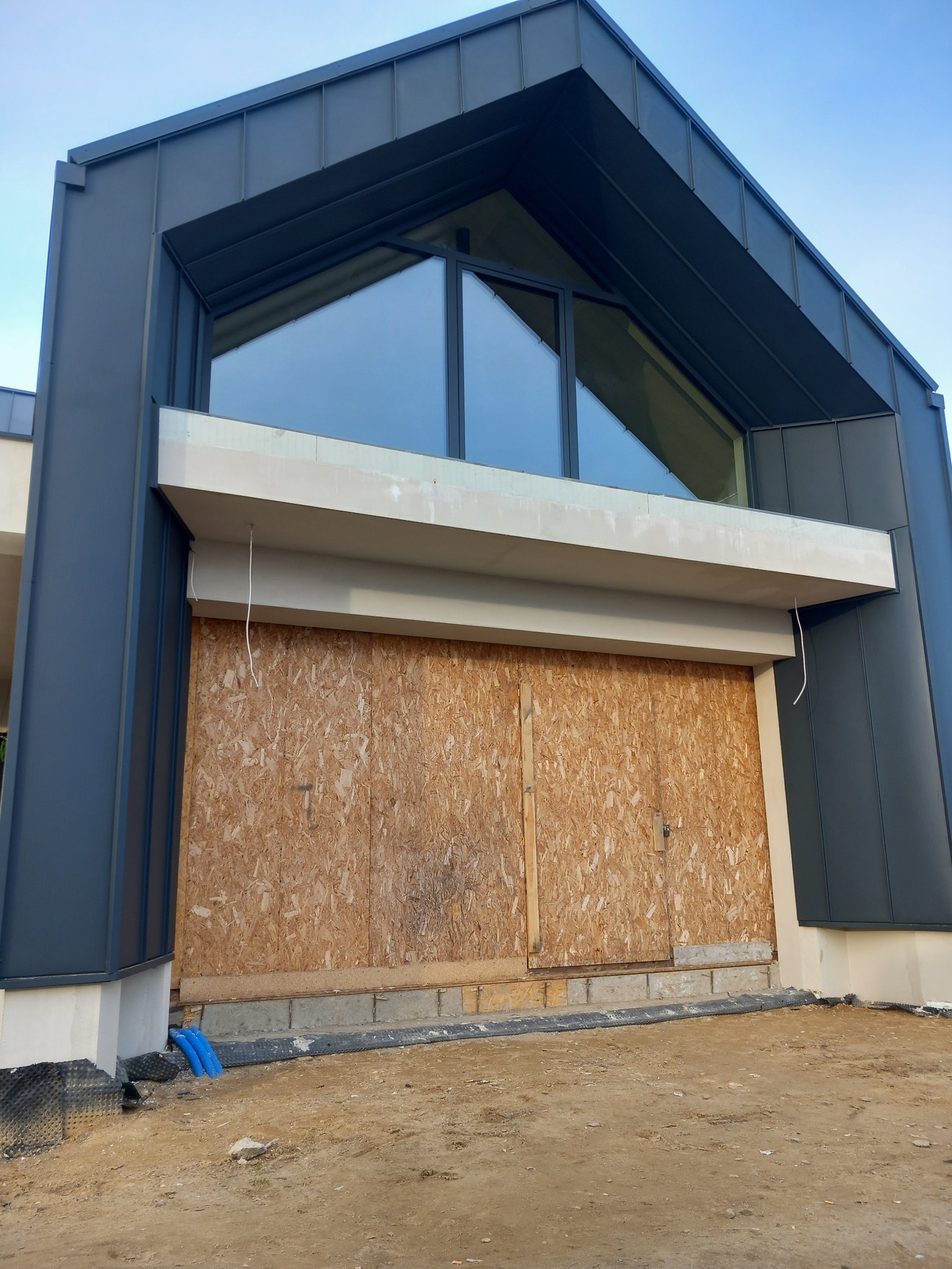 Nowoczesny budynek w trakcie budowy z szarym metalowym wykończeniem dachu i ścian, okno w kształcie trójkąta, wejście tymczasowo zabezpieczone płytami OSB.